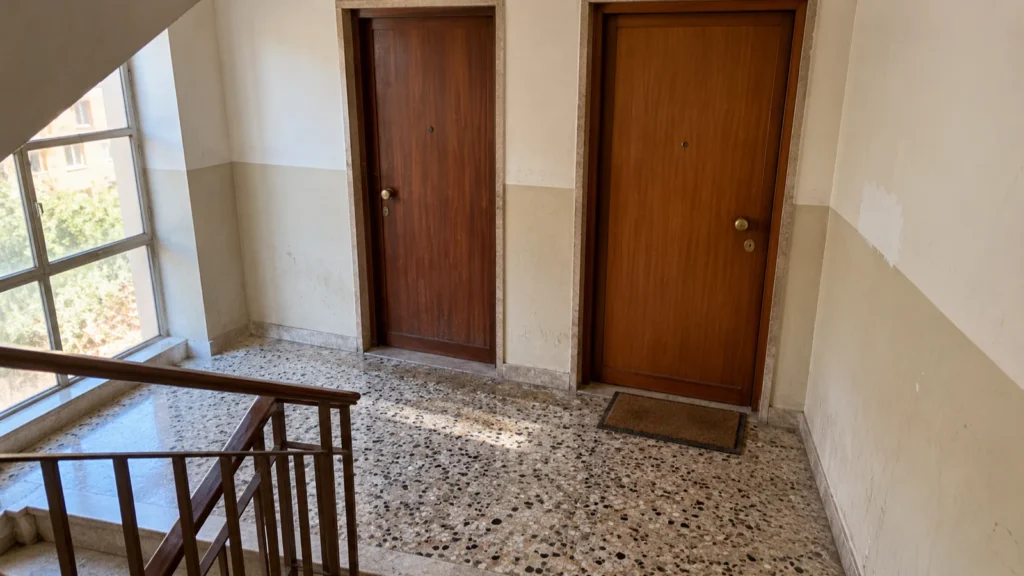 Pianerottolo di un condominio con due porte d'ingresso in legno affiancate, pavimento alla veneziana e finestra sulla scala